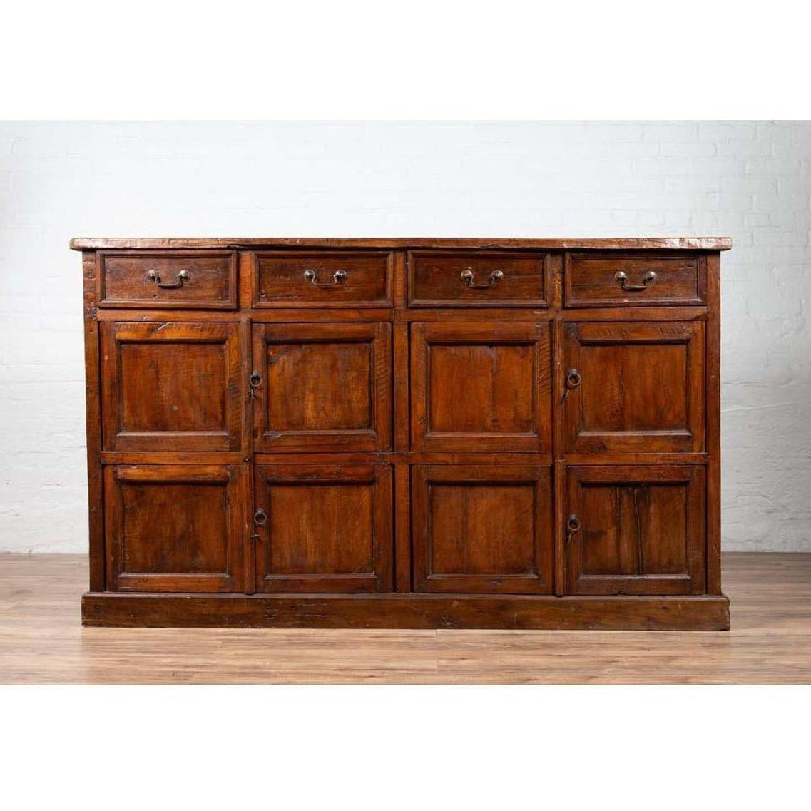 Brown teak wood cabinet with four drawers and four double doors, set against a white brick wall indoors.