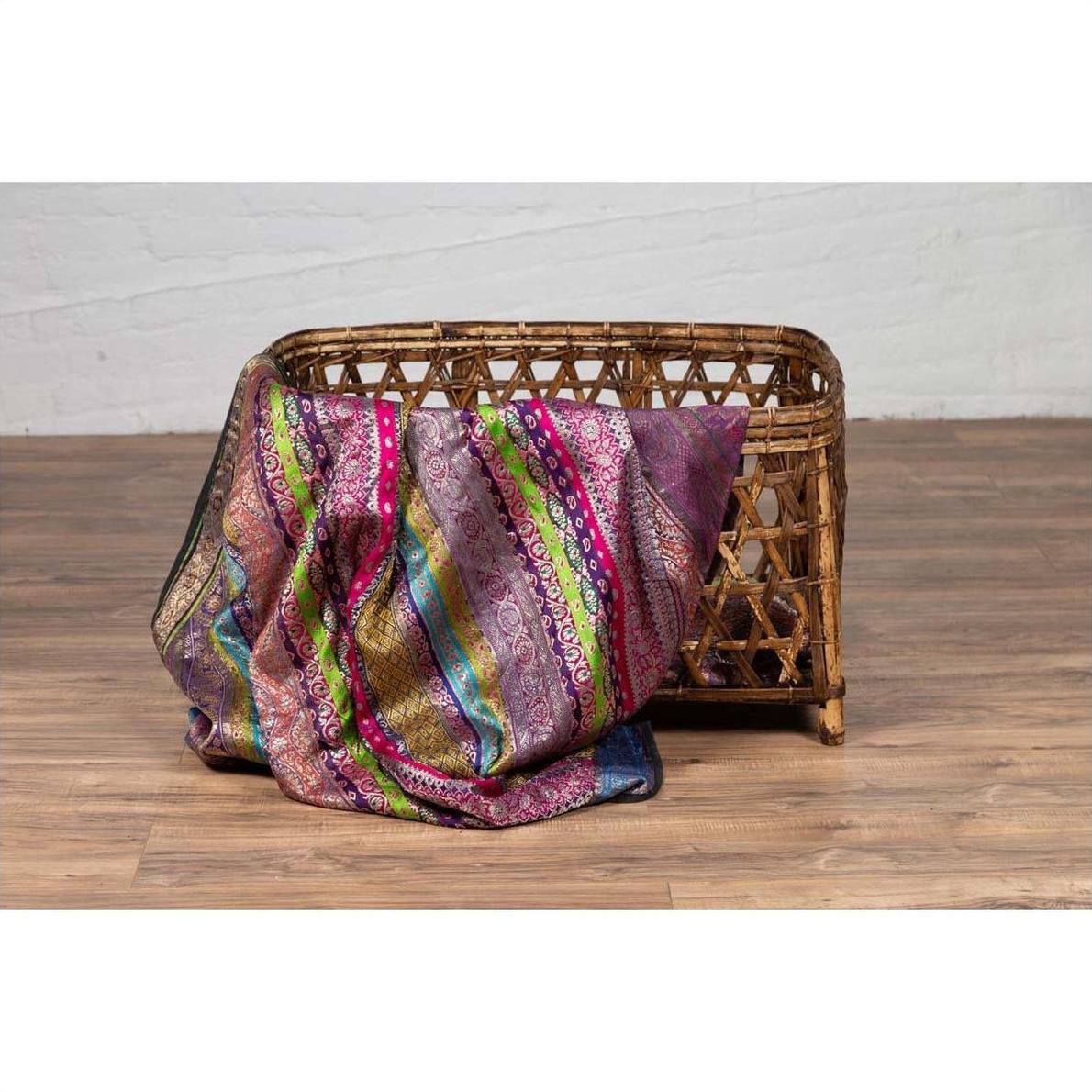 Colorful patterned fabric on a large vintage Thai bamboo fretwork basket, circa 1950, on a wooden floor.