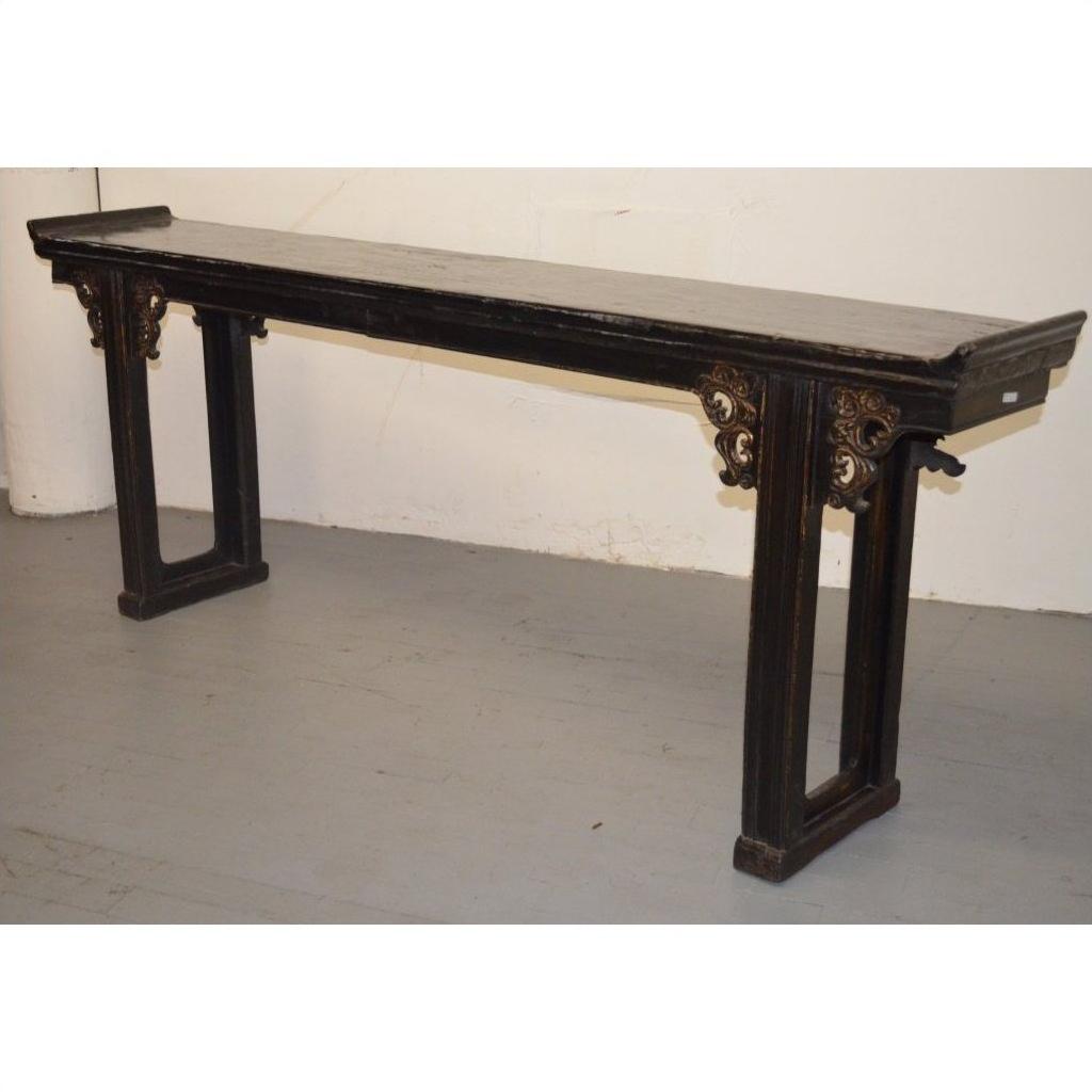 Dark wood antique Chinese altar table with carved spandrels, positioned against a plain white wall and grey floor.
