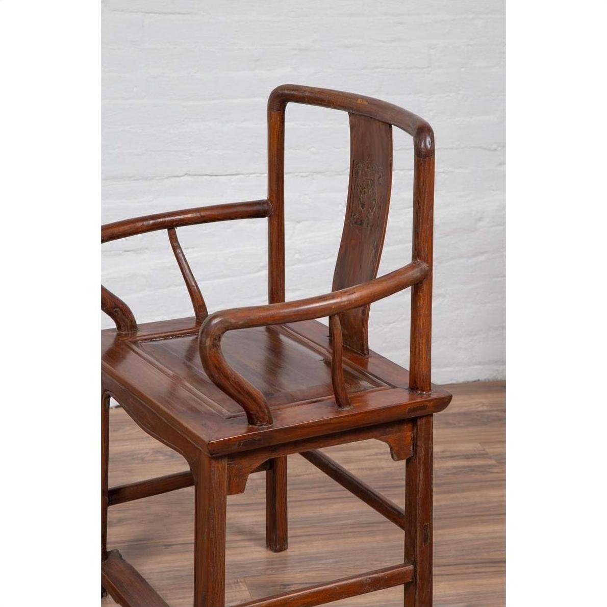 Brown wooden Ming Dynasty style wedding chair with carved medallion and curving arms next to white brick wall indoors.