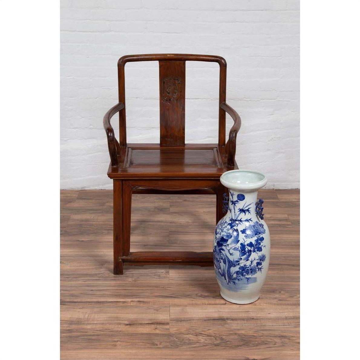 Brown wooden Ming Dynasty style wedding chair next to blue and white floral ceramic vase on wooden floor indoors.