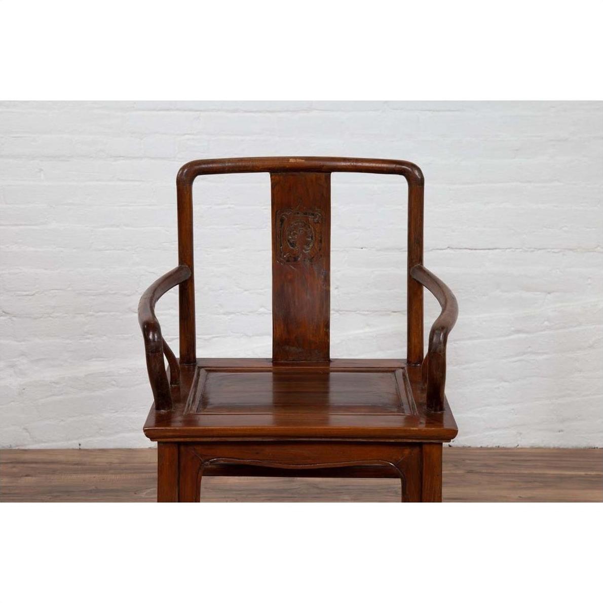 Brown wooden chair with curving arms and carved medallion, placed indoors against a white brick wall.