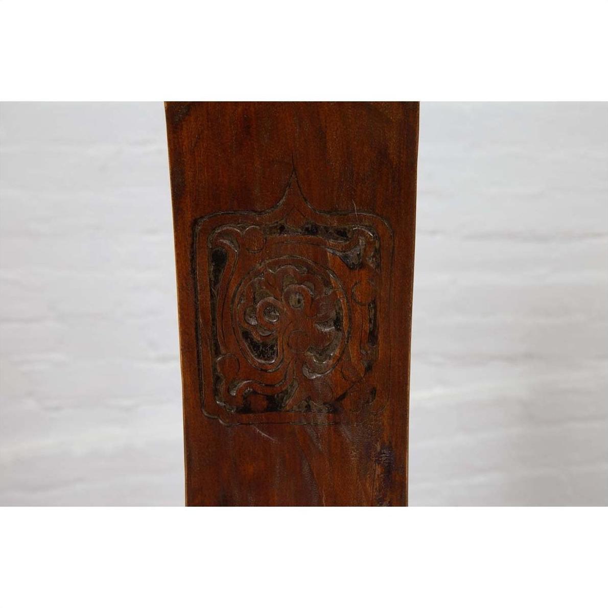Brown wood chair close-up with carved medallion and curved arms, Ming Dynasty style, blurred background visible.