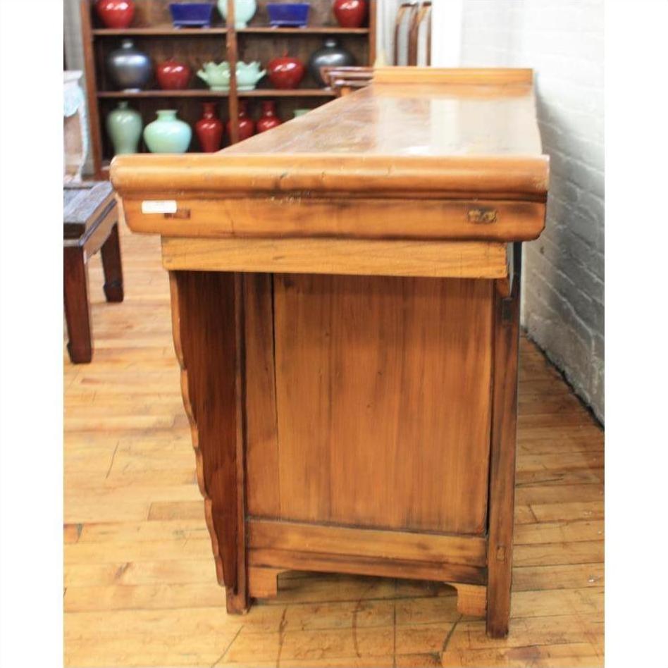 Natural wood altar coffer with scalloped edges and brass hardware, placed near shelves of colorful vases.