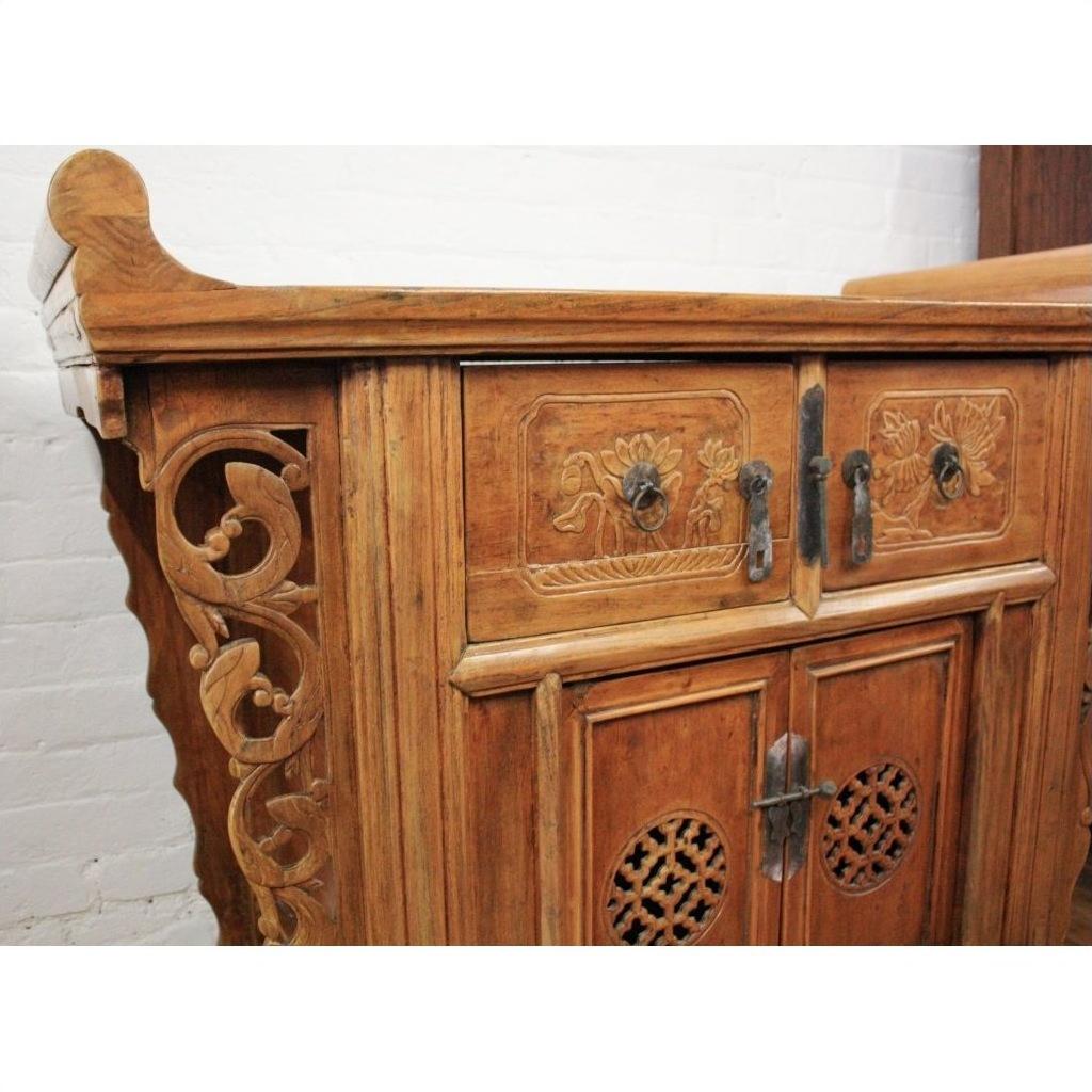 Natural elmwood altar cabinet with floral carvings, ornate details, and metal handles against a white brick wall.