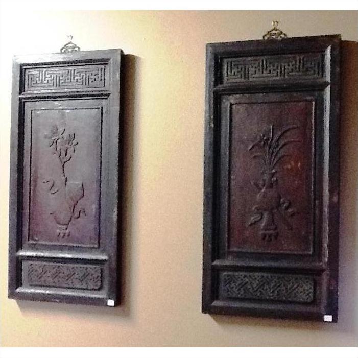 Brown wooden antique Chinese panels with simple carvings and floral designs hanging side by side on a beige wall.