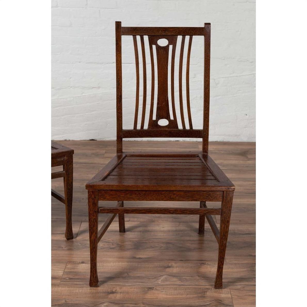 Brown wood Indonesian side chairs with pierced backrests, on wooden floor near a white brick wall indoors.