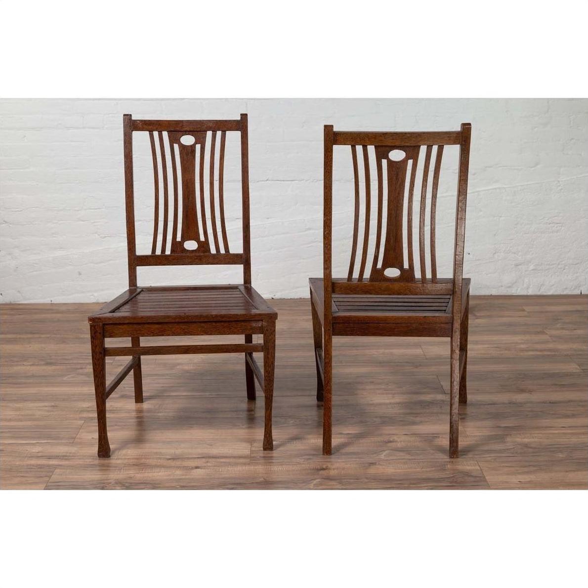 Brown wooden vintage Indonesian side chairs with pierced splats on a wooden floor by a white brick wall indoors.