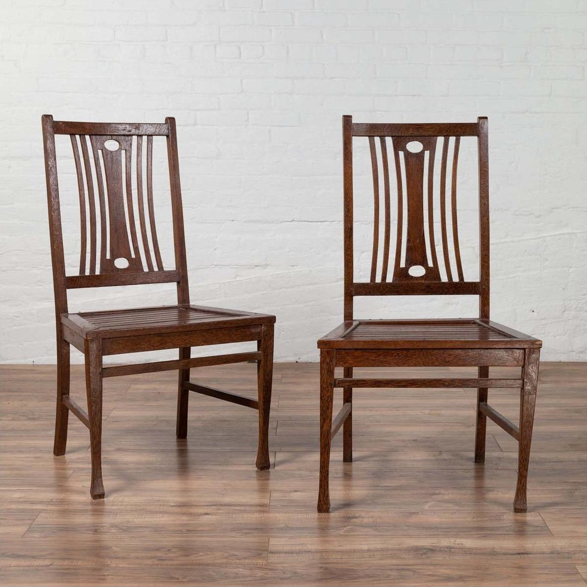 Brown wooden Indonesian side chairs with pierced backs, mid-century style, indoors by white brick wall and wood floor.