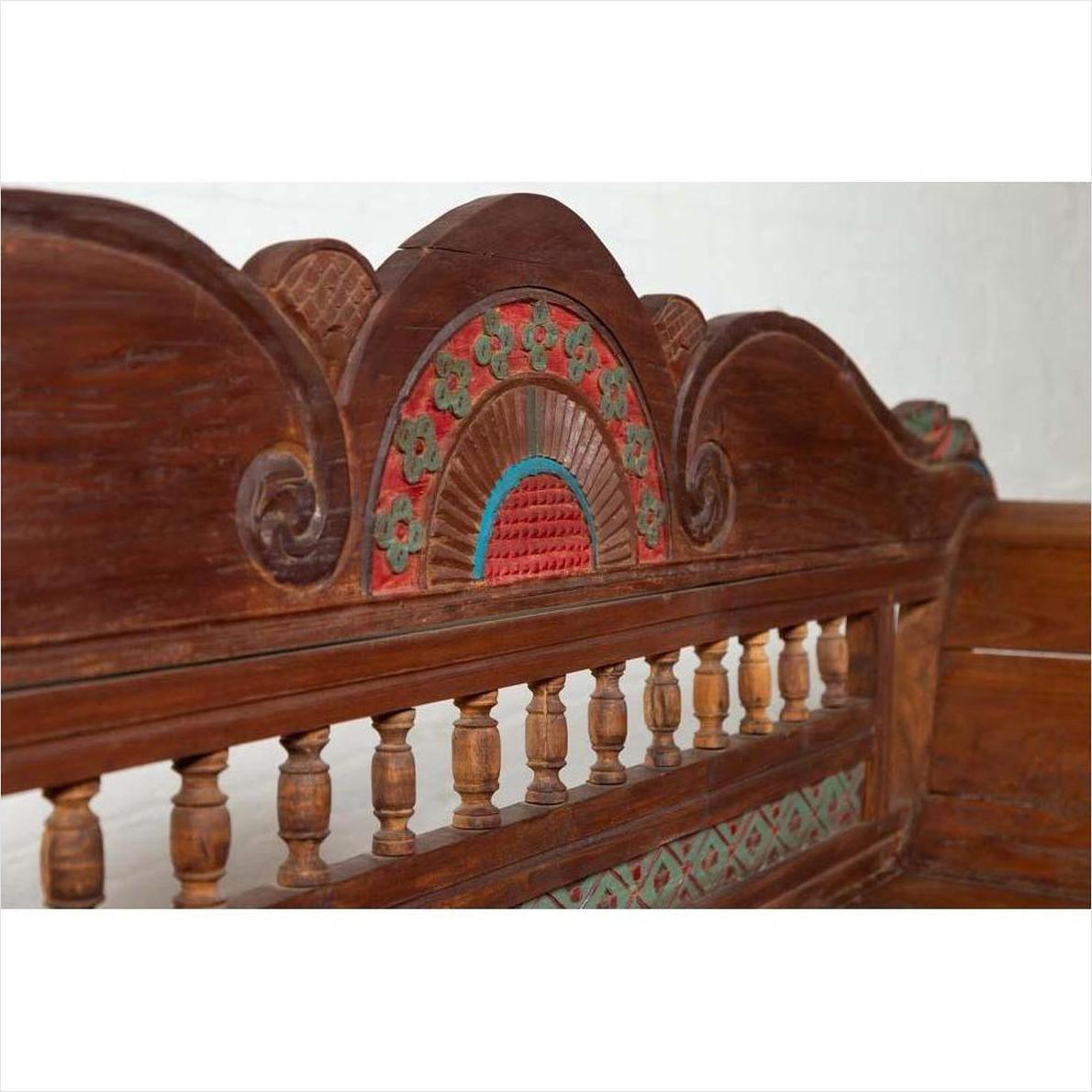 Close-up of brown teak wood settee with polychrome decor and out-scrolling arms on the backrest, indoor setting.