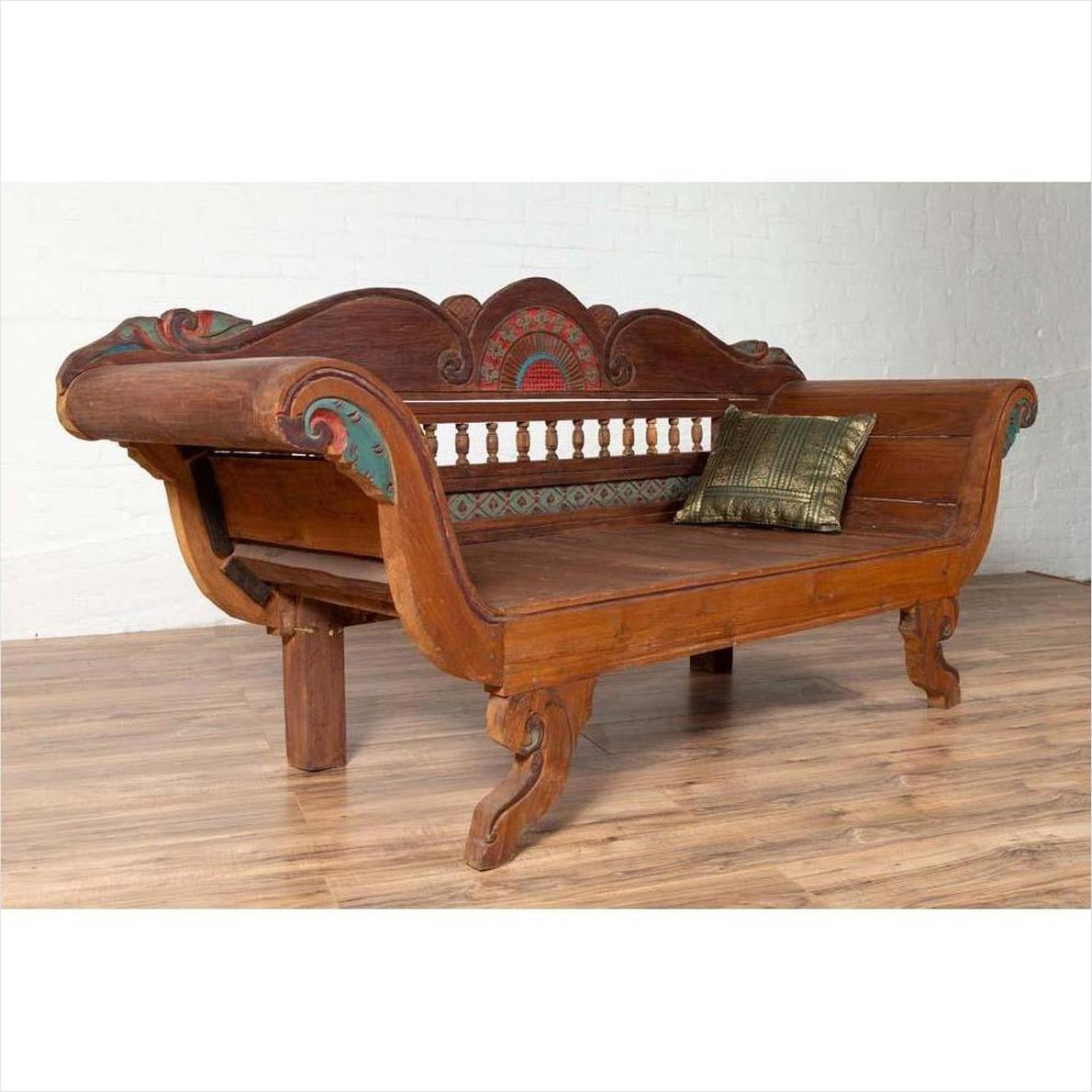 Brown teak wood settee with multicolored decorative details, out-scrolling arms, and gold pillow on wooden floor.