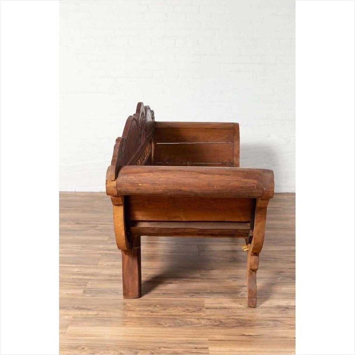 Brown teak wood settee with polychrome details and out-scrolling arms on a wooden floor, side view.