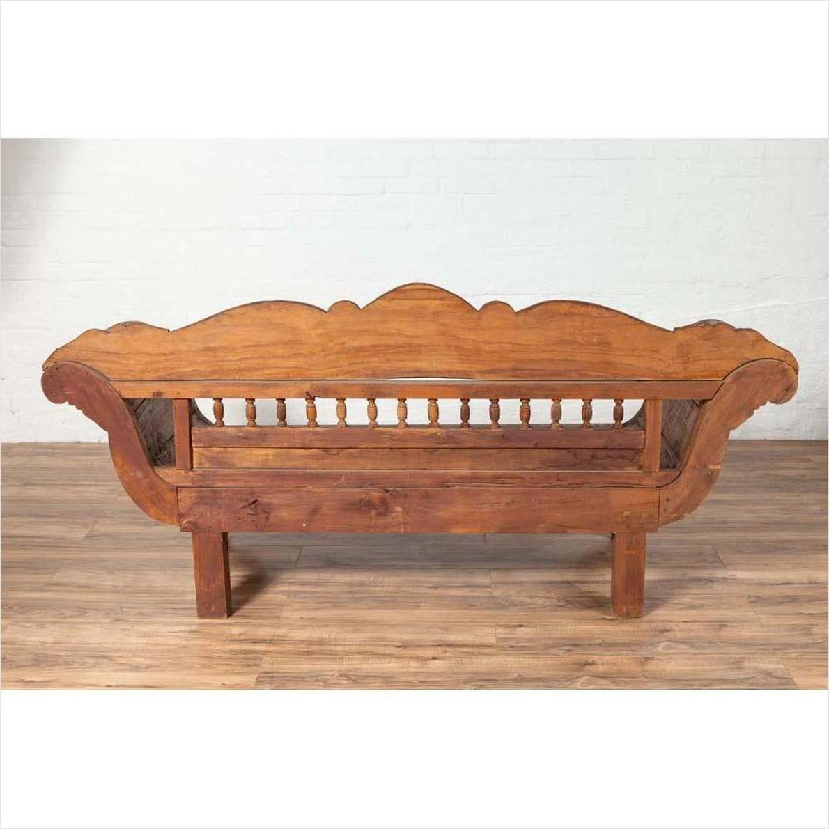 Brown teak wood settee with polychrome décor and out-scrolling arms on wooden floor against white wall indoors.