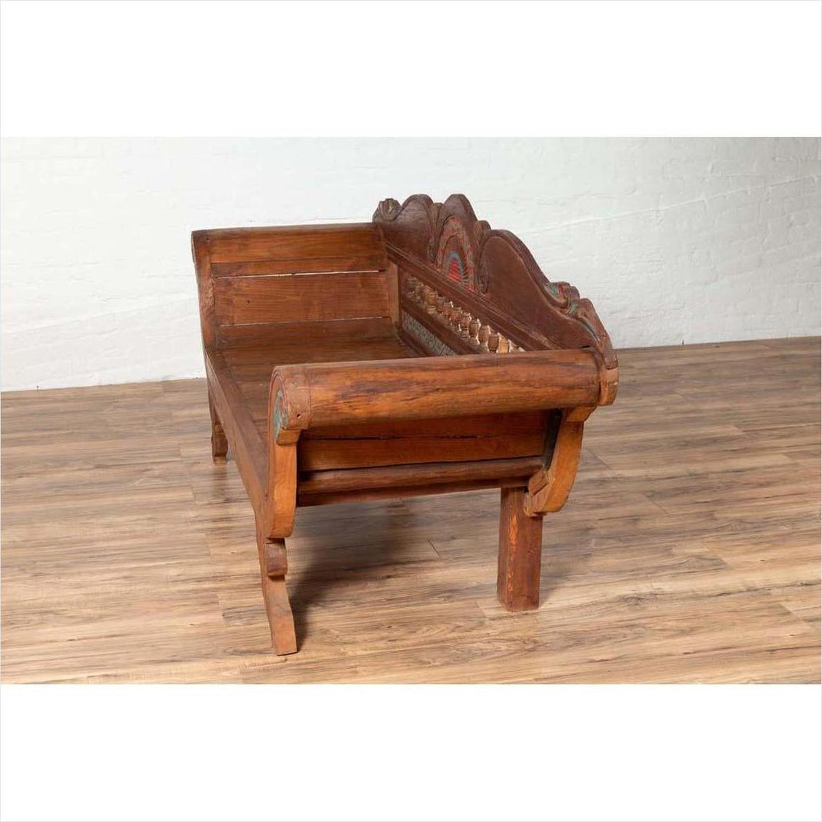 Brown teak wood settee with polychrome decor and out-scrolling arms on wooden floor against white brick wall.