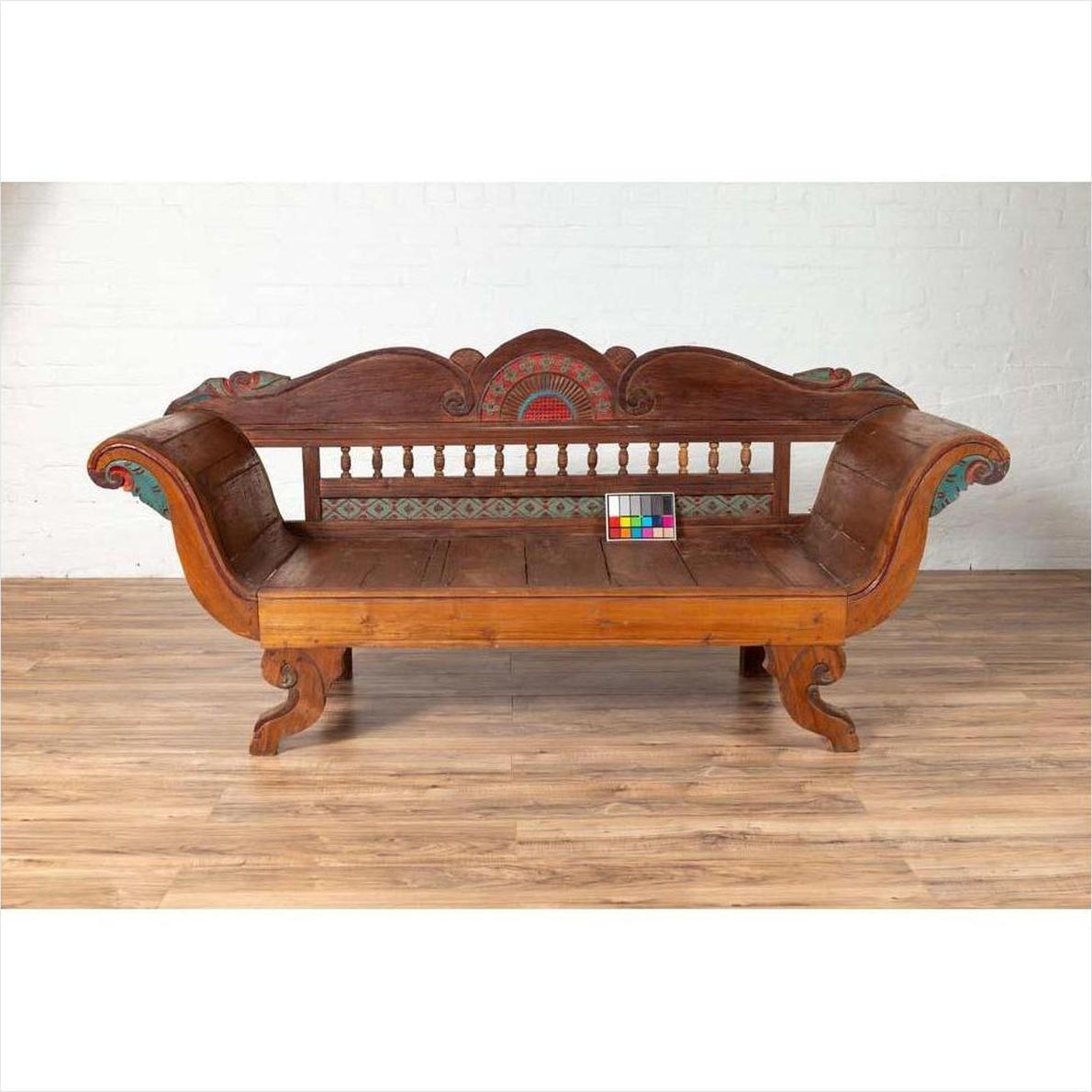 Brown teak wood settee with polychrome décor and out-scrolling arms on wood floor against white brick wall indoors.