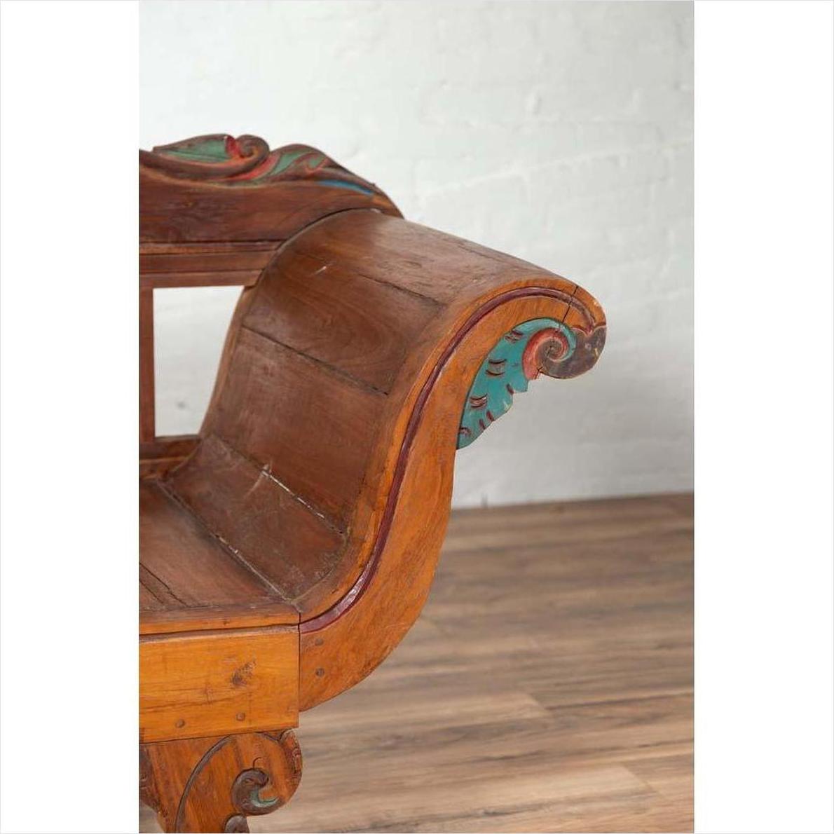 Brown teak wood settee with colorful polychrome décor and curved arms, placed on a wooden floor indoors.