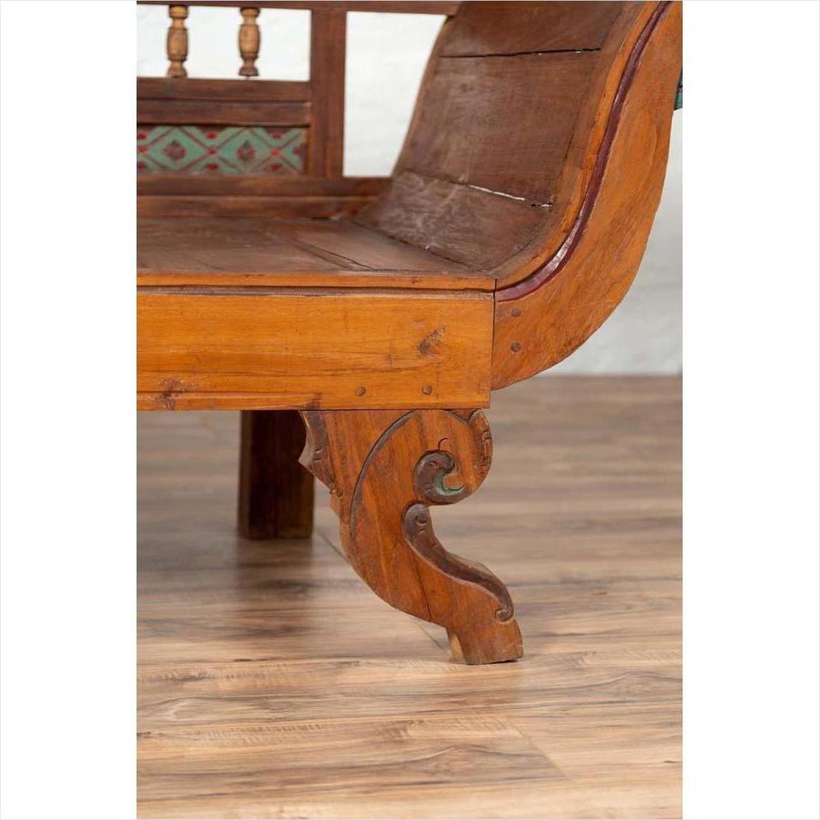 Close-up of brown teak wood settee with multicolor décor and out-scrolling arms on light wood floor indoors.