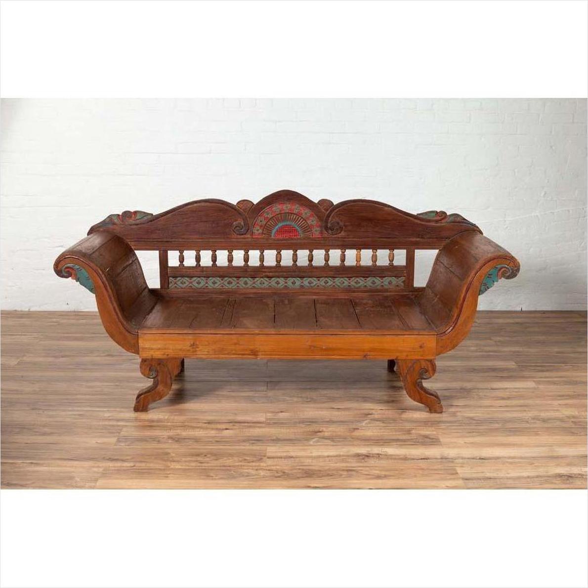Brown teak wood settee with colorful polychrome décor and out-scrolling arms, placed on a wooden floor indoors.