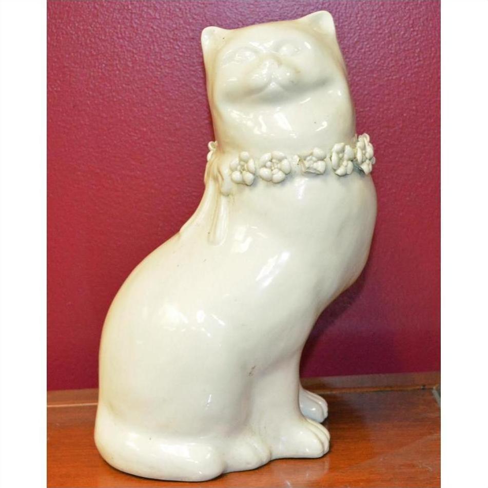 White porcelain cat figurine with floral collar, head turned to side, on wooden surface against red wall.