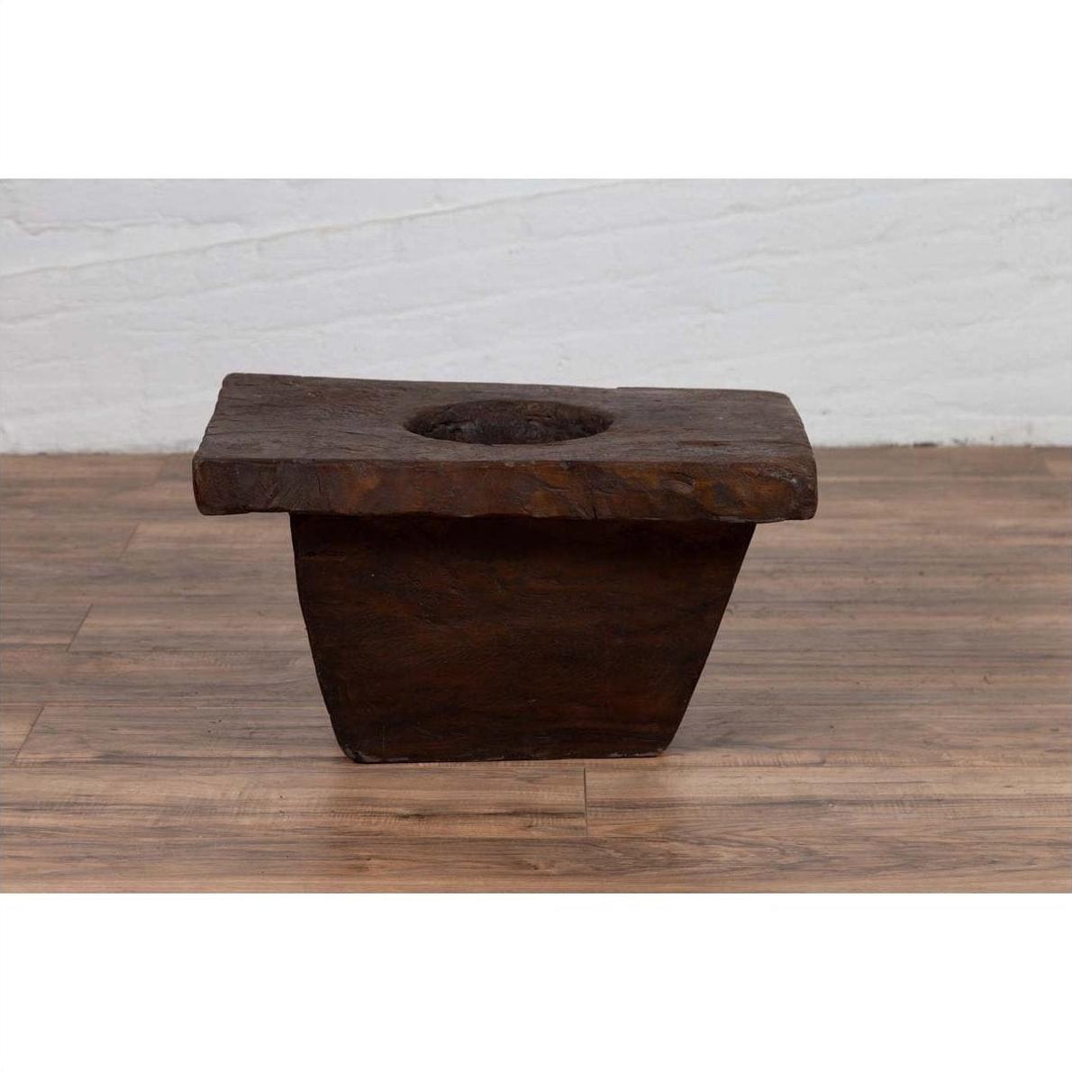 Brown wooden planter, antique Indonesian style, placed on a wood floor next to a white brick wall indoors.