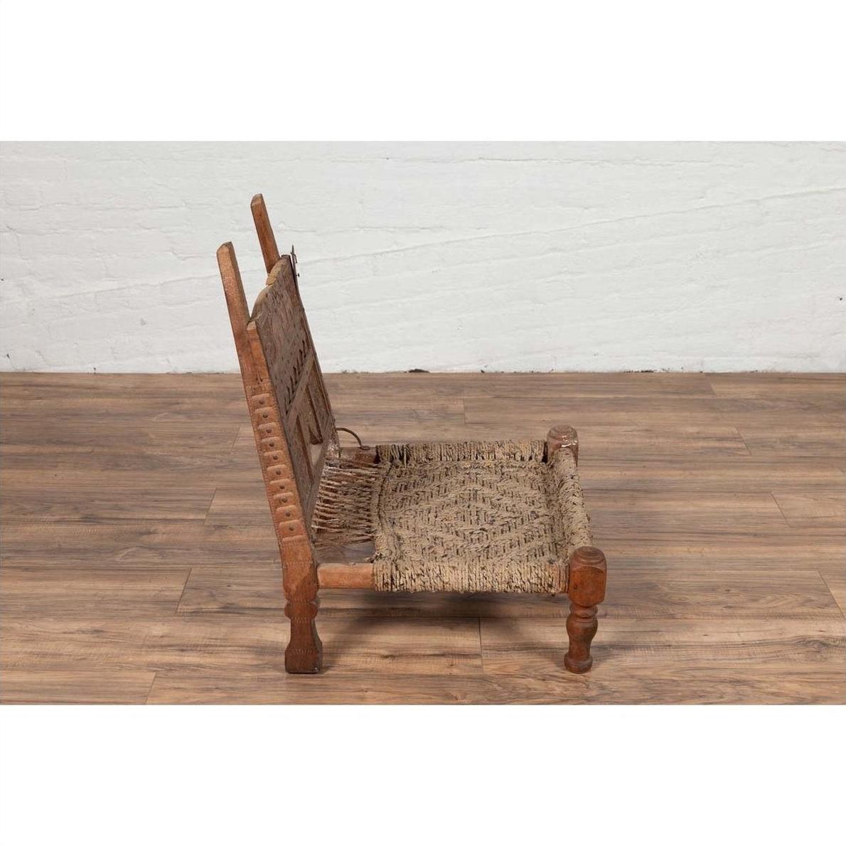 Brown wooden low chair with rope seat, rustic and weathered, on wooden floor by a white brick wall indoors.