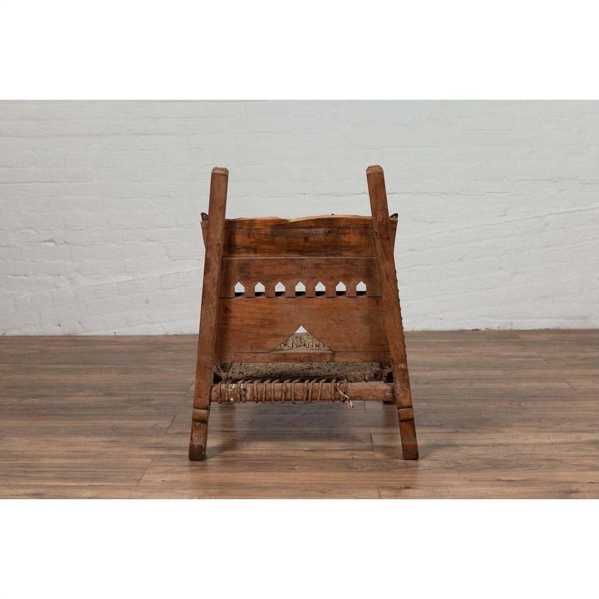 Brown wood and rope low chair with weathered finish on a wooden floor next to a white brick wall indoors.