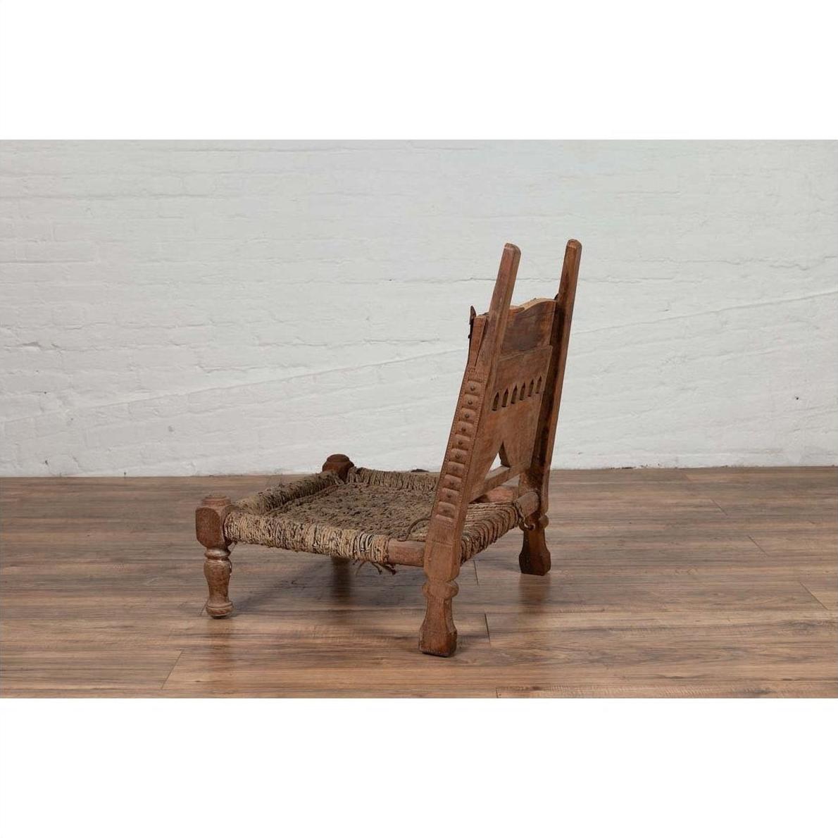 Brown wooden low chair with rope seat and weathered finish on a wooden floor next to a white brick wall indoors.