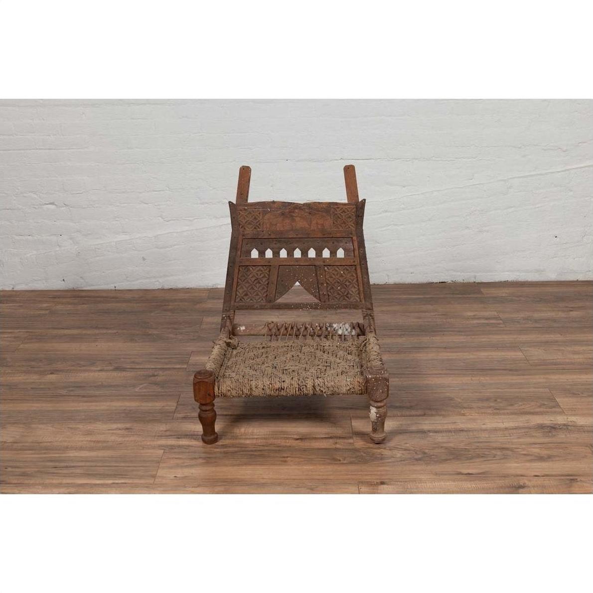 Brown wood and rope low Indian chair with weathered finish on wooden floor near a white wall indoors.