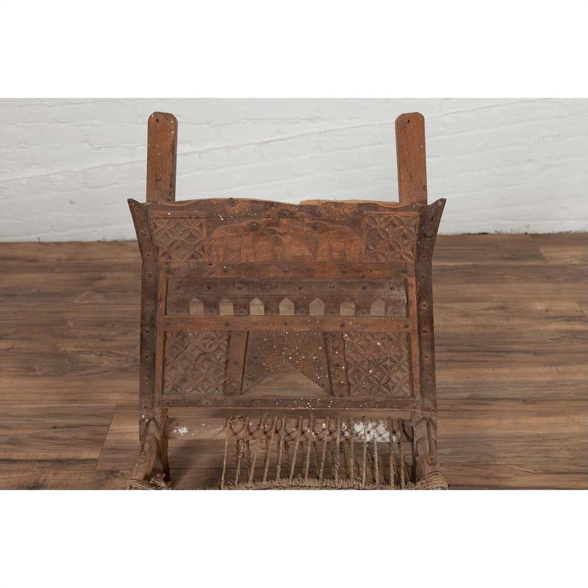 Brown wooden low chair with rope seat, weathered finish, placed on a wooden floor beside a white brick wall indoors.