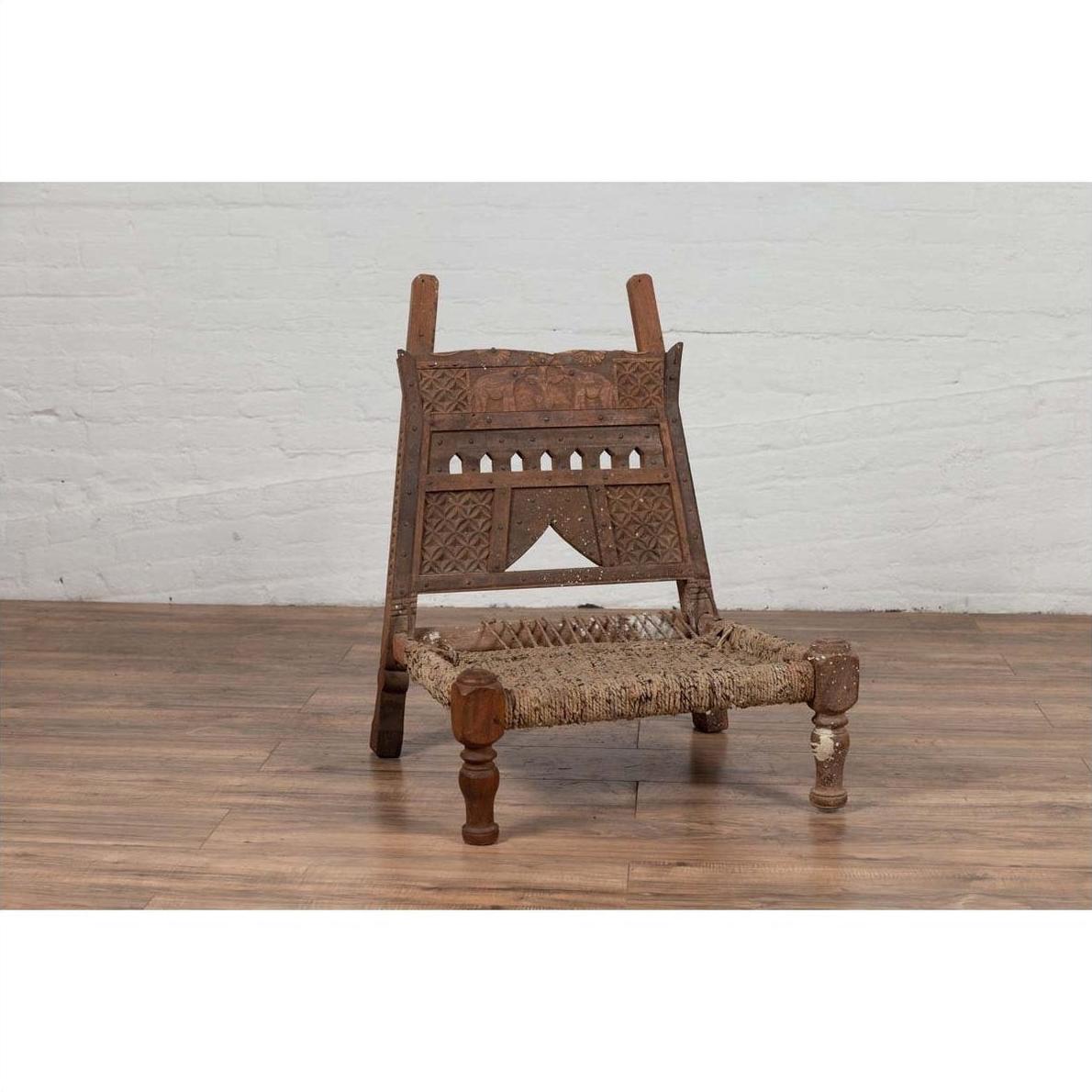 Brown wooden low chair with rope seat, weathered look, on wooden floor next to white brick wall indoors.