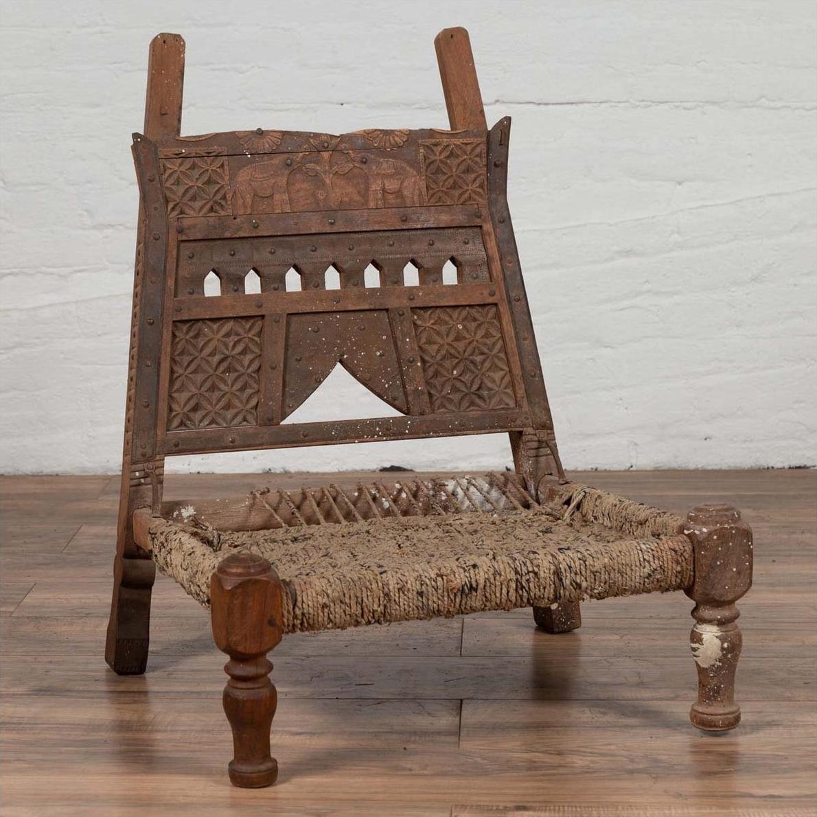 Brown wooden low chair with rope seat and weathered finish on a wooden floor, next to a white brick wall indoors.