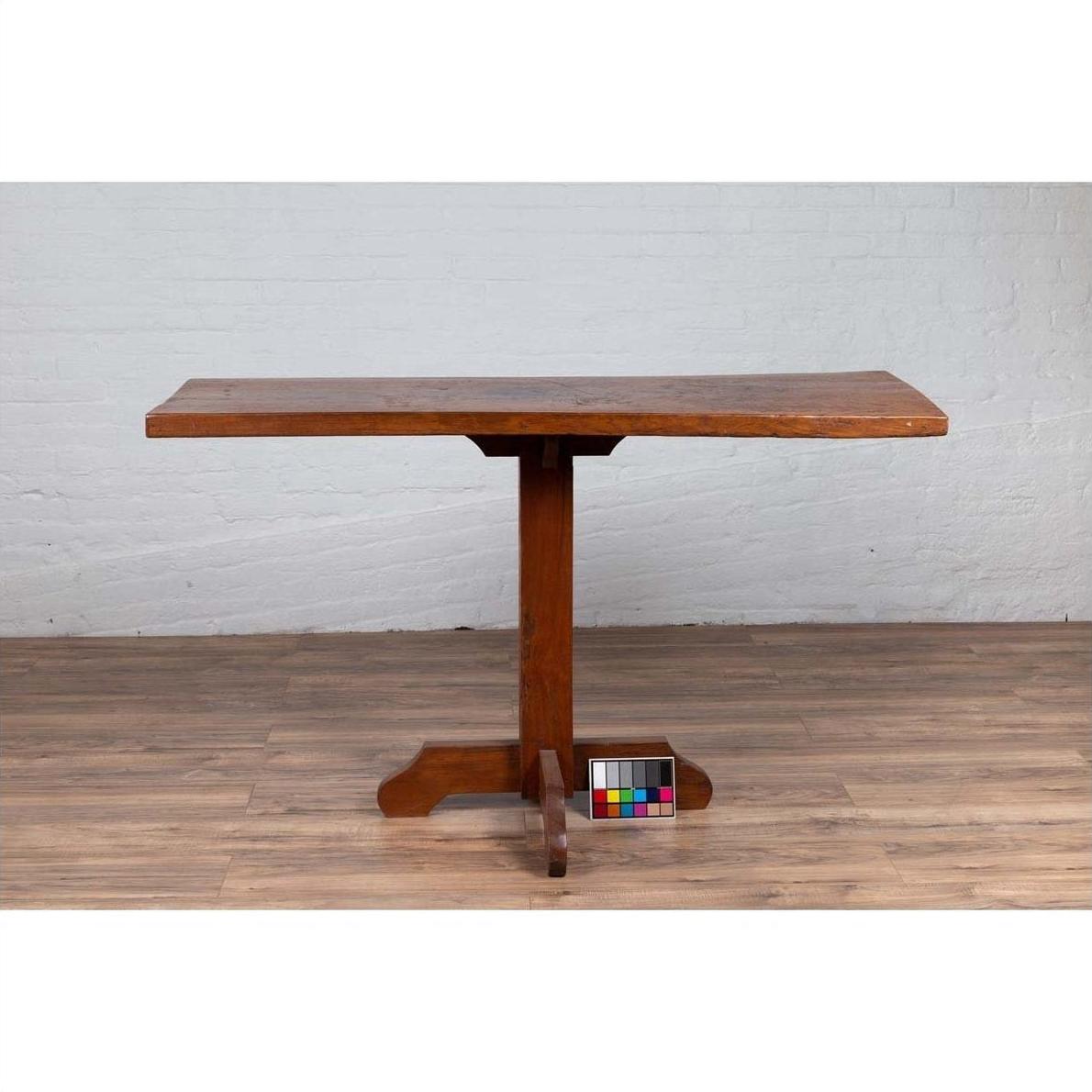 Brown wooden Indonesian console table with single plank top and pedestal base on wooden floor, front view.