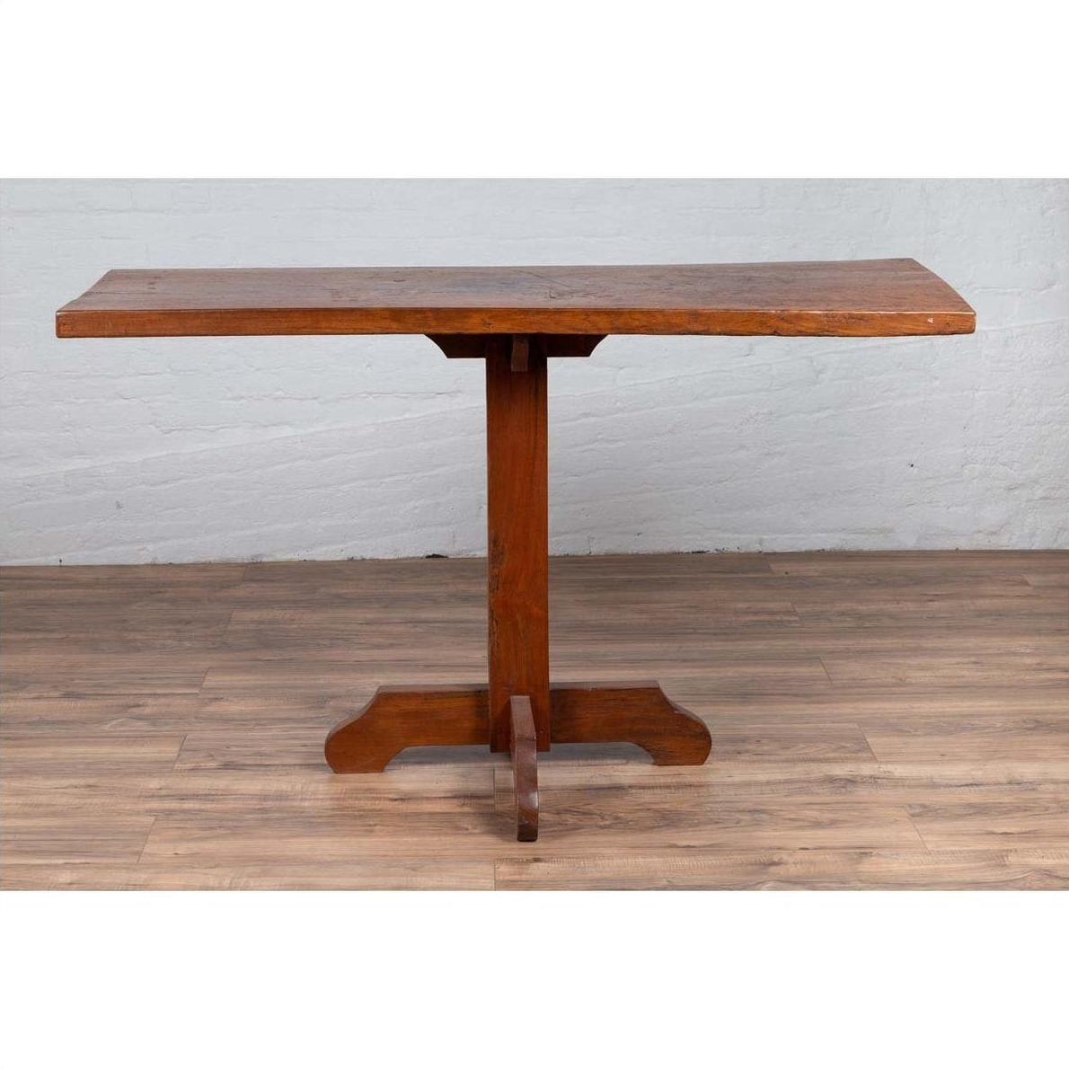 Brown wooden Indonesian console table with single plank top and pedestal base on wood floor against white brick wall.