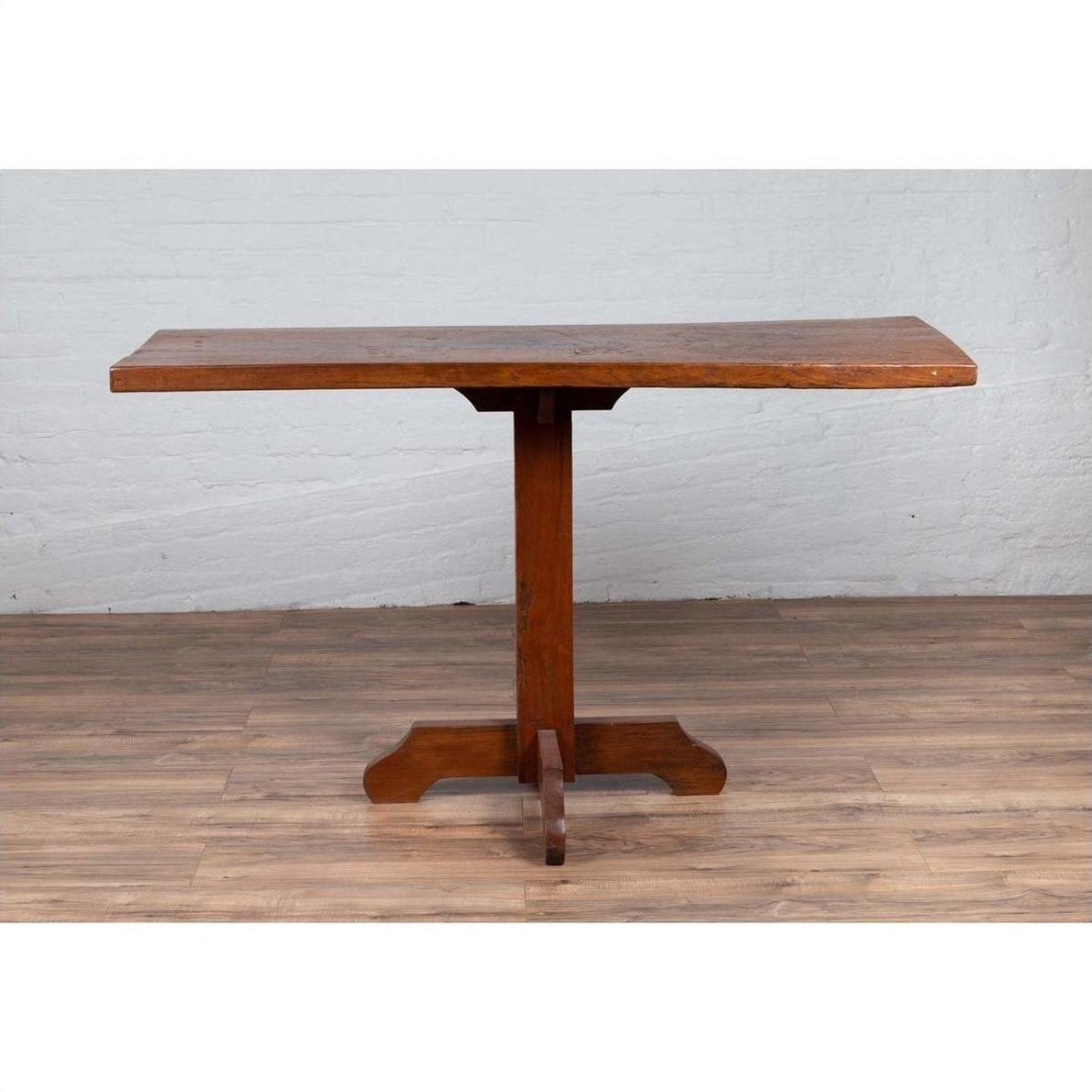 Natural wood Indonesian console table with single plank top and pedestal base on wooden floor against white brick wall.