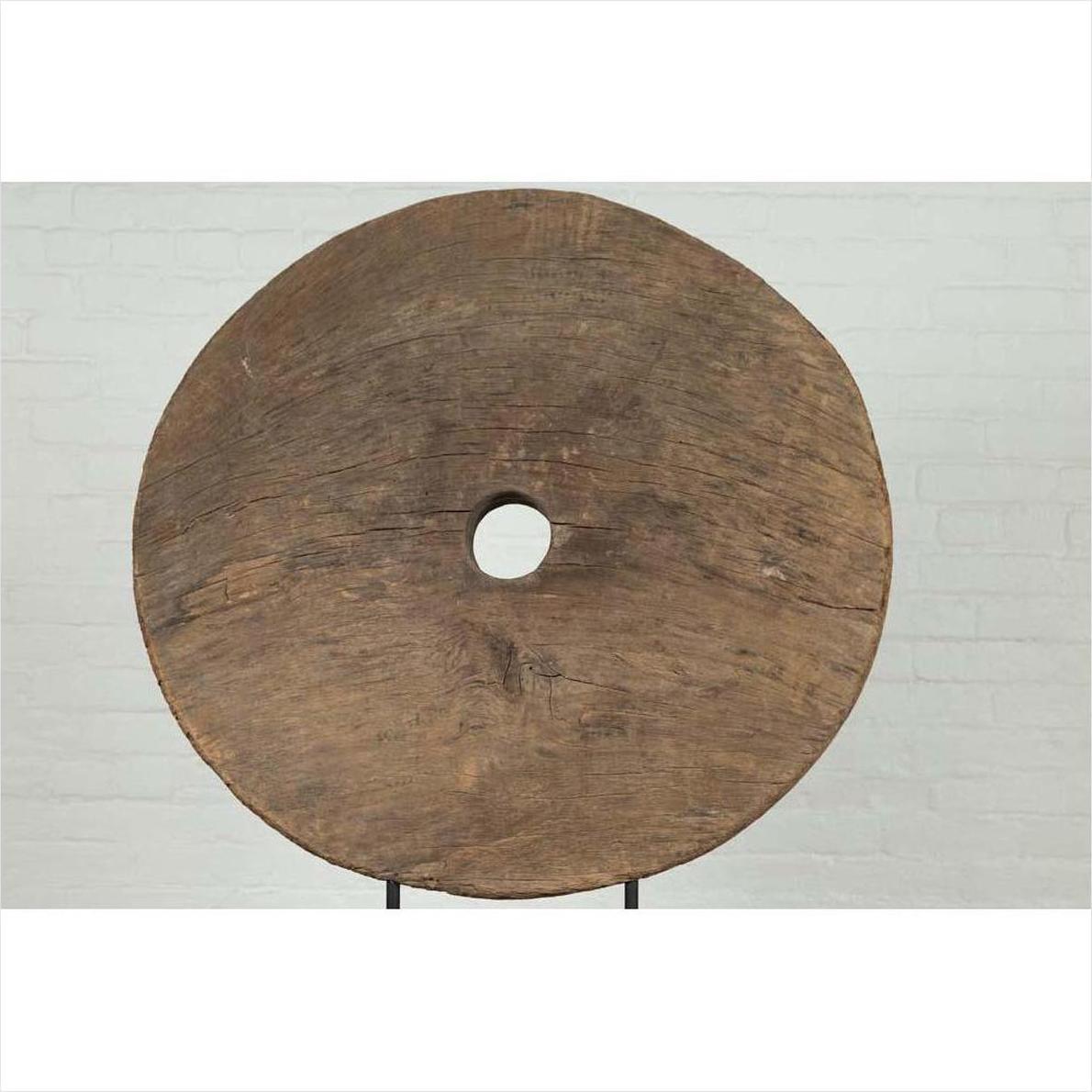 Brown wooden ox cart wheel on black base, displayed indoors in front of a light brick wall.