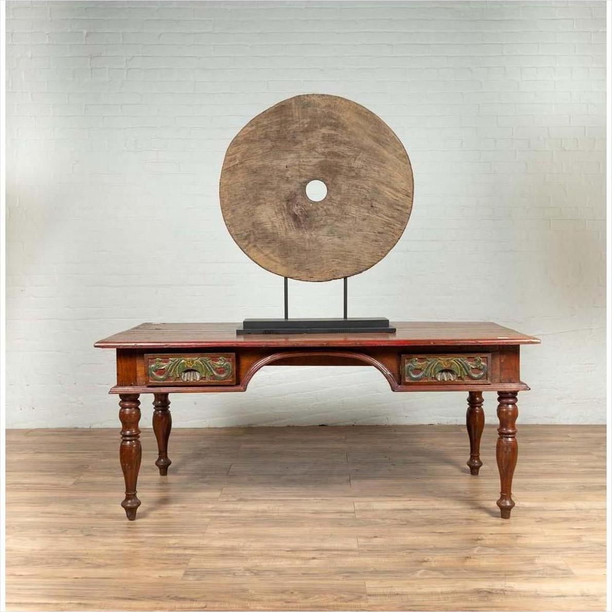 Brown wooden ox cart wheel on black painted base, rustic style, displayed behind an antique table indoors.
