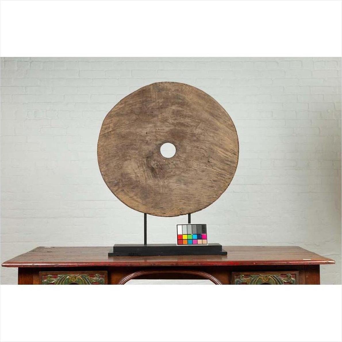 Brown wooden ox cart wheel from Thailand on black base, placed on a wooden table with white brick wall background.