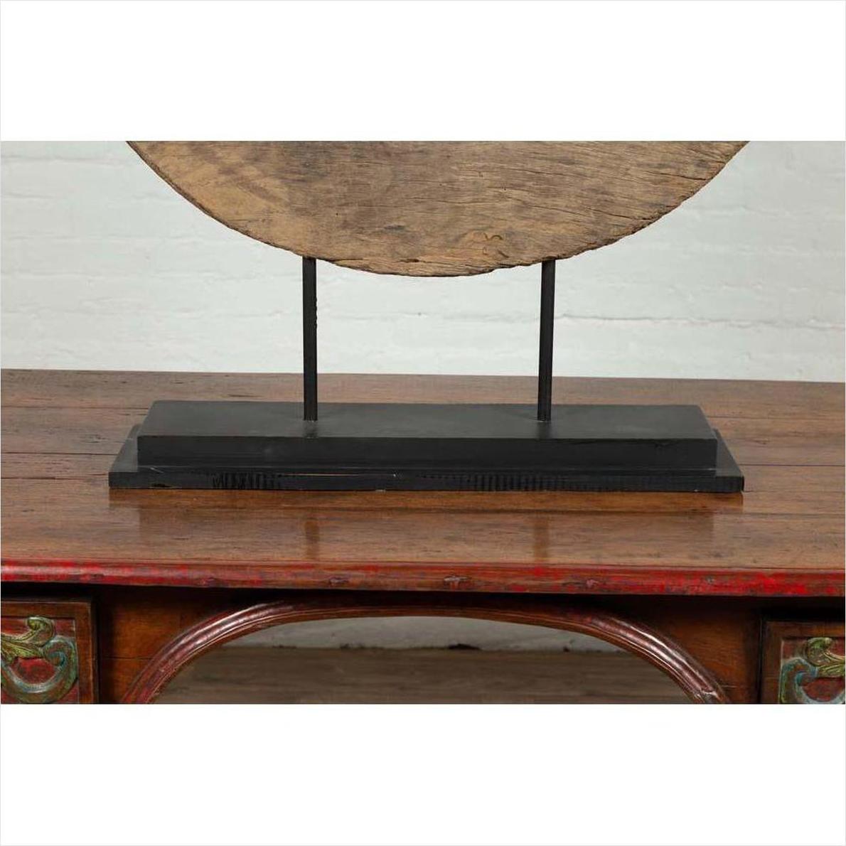 Brown wooden ox cart wheel from Thailand on black base, displayed on polished carved wooden table indoors.