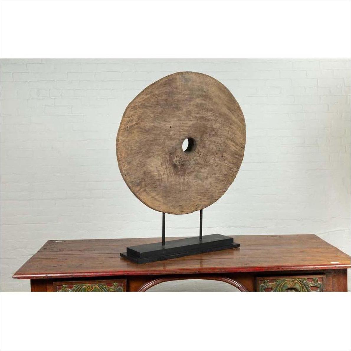Brown wooden ox cart wheel on black base, displayed on a table with white brick wall background.
