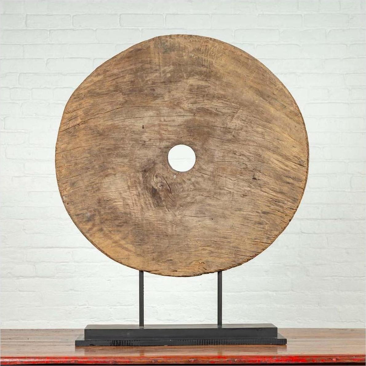 Brown wooden ox cart wheel from Thailand on a black base, displayed indoors against a white brick wall.