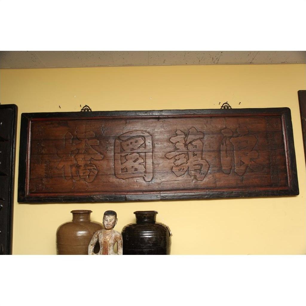 Black Chinese calligraphy signboard on yellow wall, above ceramic pottery and ornate figurine, indoor setting.