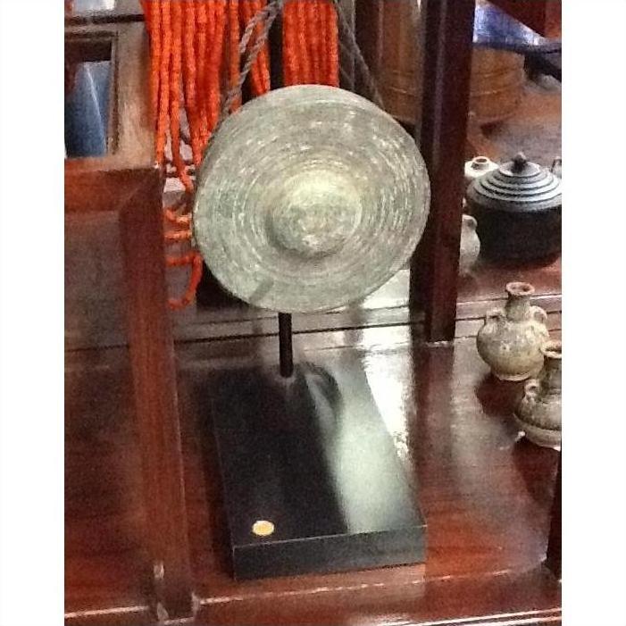 Close-up of a small metal gong on a black stand, with pottery and beads in the background.