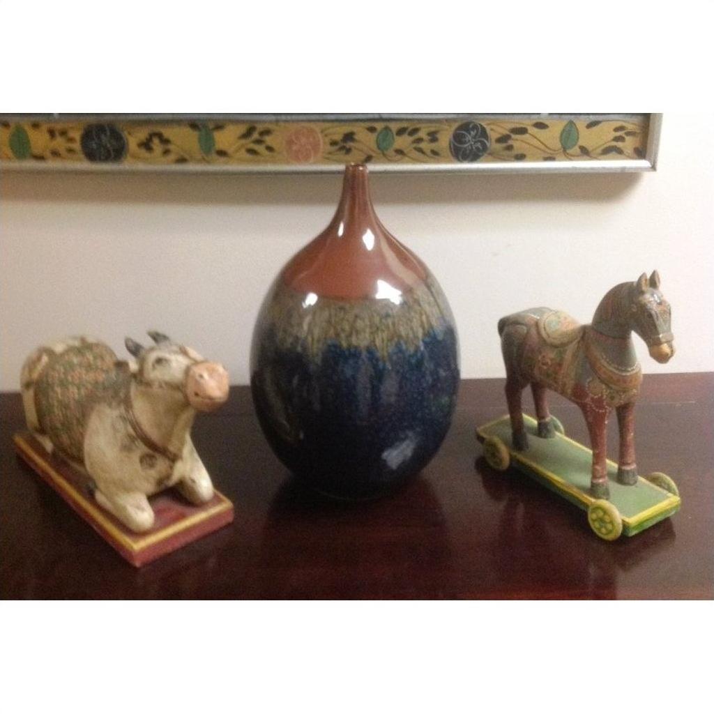 Close-up of a brown and blue ceramic Prem vase on a wooden surface, between wheeled cow and horse figurines.