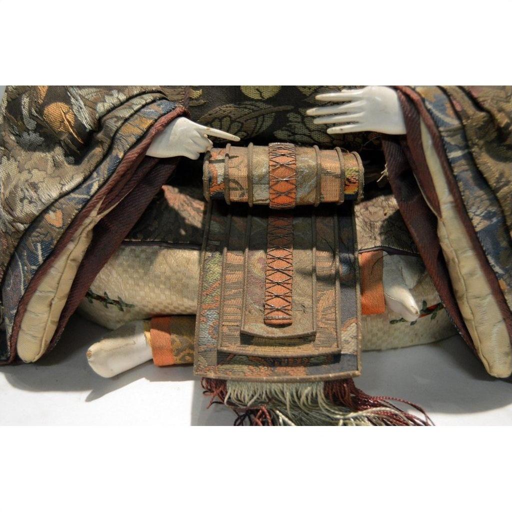 Close-up of a Taisho Samurai doll in patterned fabric robes holding a small decorative item with both hands.