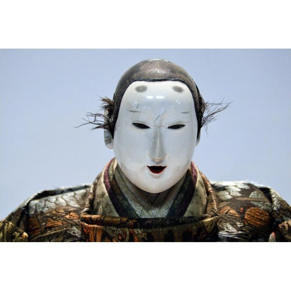 Close-up of a Taisho samurai doll with a white Noh mask, black hair tufts, and ornate patterned clothing.