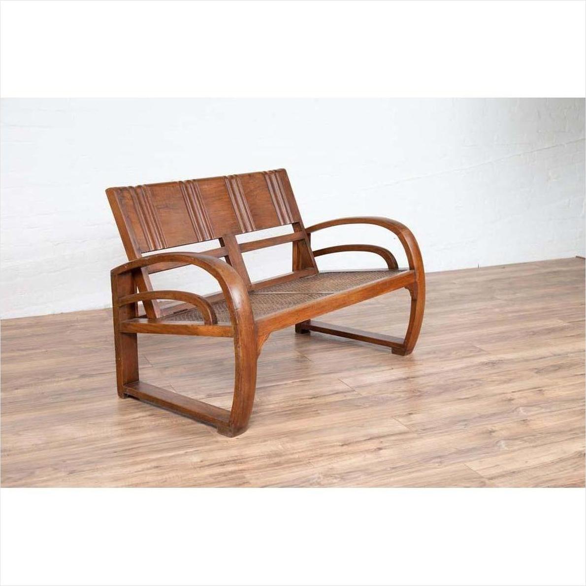 Brown teak wood settee with folding back, looping arms, and cane seat on light wooden floor indoors against white wall.