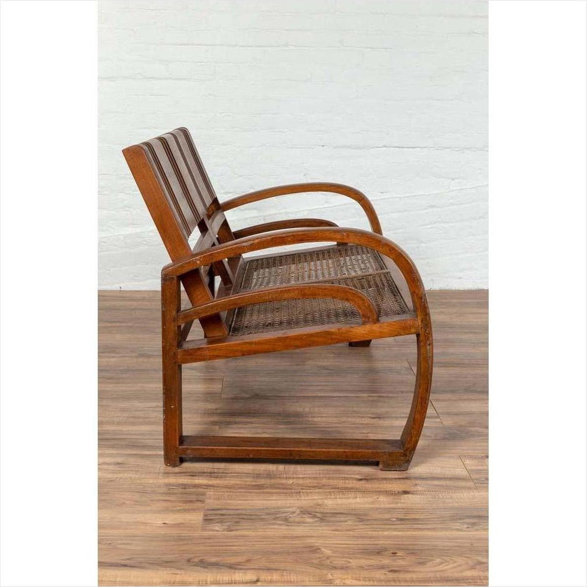 Brown teak wood settee with folding back, looping arms, and cane seat on wooden floor, side view.