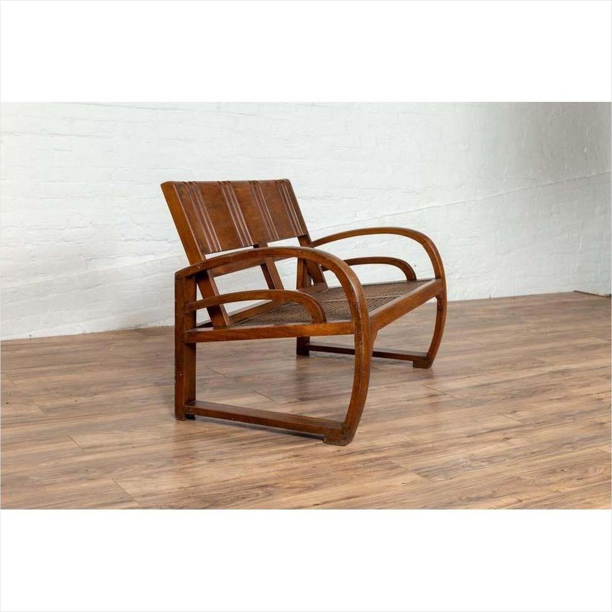 Brown teak wood settee with folding back, looping arms, and cane seat on wooden floor, white brick wall background.