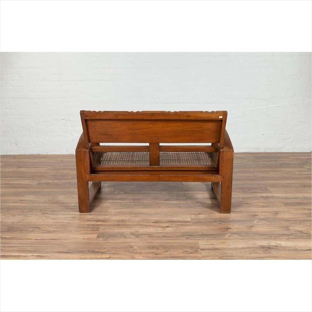 Brown teak wood settee with folding back, looping arms, and cane seat against a white brick wall indoors.