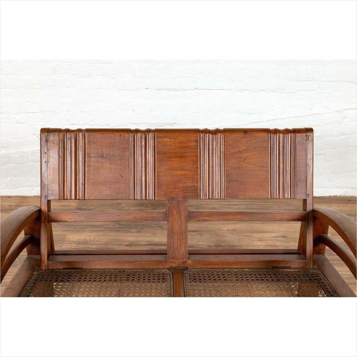 Brown teak wood settee with folding back, looping arms, and cane seat against a white brick wall indoors.